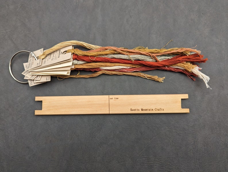 Pode incluir: Um tear de madeira com uma etiqueta que diz "Scotts Mountain Crafts" e um conjunto de fio de bordado em v&aacute;rios tons de marrom, laranja e vermelho.