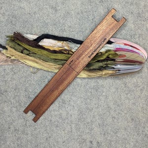 May include: A wooden embroidery floss organizer with the text "Scents Mountain Crafts" printed on it. The organizer has two slots for holding floss and a ruler marked in inches. The organizer is lying on a gray surface with a bundle of embroidery floss in various colors.