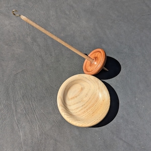 May include: A wooden spinning wheel with a spindle and a separate wooden bowl. The spinning wheel is made of light brown wood with a darker brown center. The bowl is made of light brown wood with a grain pattern.