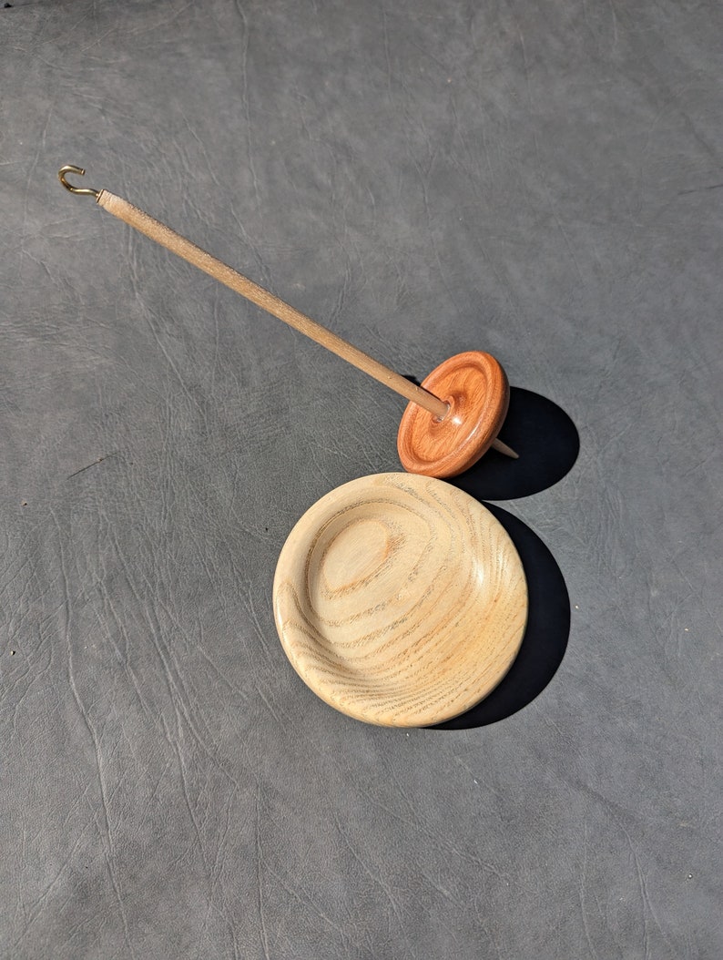 May include: A wooden spinning wheel with a spindle and a separate wooden bowl. The spinning wheel is made of light brown wood with a darker brown center. The bowl is made of light brown wood with a grain pattern.