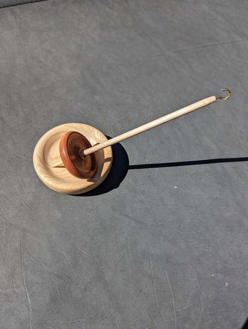 May include: A wooden spinning wheel with a natural wood finish and a brown wooden spindle. The wheel is sitting on a wooden base.