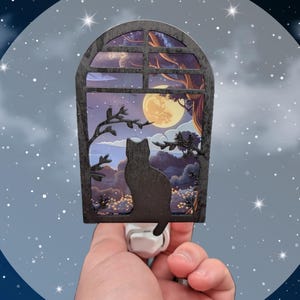 May include: A black cat silhouette sits in a window looking out at a full moon and starry night. The window is made of wood and has a decorative design. The nightlight plugs into a standard outlet.