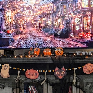 Puede incluir: Un televisor muestra una escena de Halloween con una calle con edificios iluminados y calabazas. Debajo, una guirnalda de recortes de Halloween y calabazas decorativas crean un ambiente festivo.