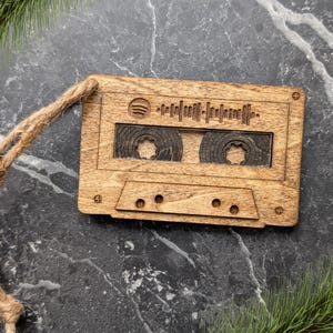 May include: Wooden cassette tape ornament with a Spotify logo and soundwave design. The ornament is made of light brown wood and has a jute twine loop for hanging. The cassette tape design includes details like the tape reels and the cassette tape holes.