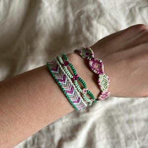 May include: Three friendship bracelets in green, pink, and white colors. The bracelets are woven with a chevron pattern and a diamond pattern.