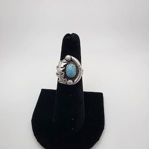 May include: A silver ring featuring a central oval turquoise stone set in a serrated bezel. The metal band is decorated with stamped leaf-like patterns and small circular accents, creating a textured appearance around the blue stone setting.