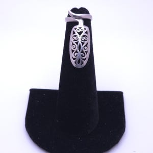 May include: A silver-coloured ring featuring an elongated, oval-shaped face with an intricate, openwork filigree pattern. The band is smooth and wraps around the finger, connecting to the decorative top section which displays symmetrical scrollwork.