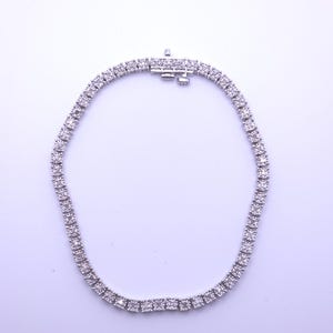 May include: A silver-toned tennis necklace featuring a continuous row of small, round, clear stones set in a metallic frame. The necklace includes a secure box clasp mechanism at the top, designed to hold the circular chain together firmly.