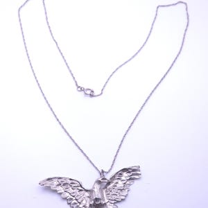 Op de afbeelding: Een zilverkleurige ketting met een hanger in de vorm van een adelaar met wijd gespreide vleugels. De adelaarshanger heeft een getextureerd, gefacetteerd oppervlak dat licht reflecteert. De hanger zit aan een dunne, fijne zilverkleurige ketting.