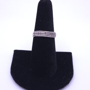 May include: A silver-toned metal ring featuring a central band with a repeating cross-hatch or X-shaped pattern, bordered by two smooth, thin horizontal lines along the top and bottom edges of the band, creating a textured appearance.