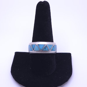 May include: A silver band ring featuring a continuous inlay of triangular turquoise stones. The metal band has a smooth, polished finish, and the vibrant blue stone segments are arranged in a repeating geometric pattern around the entire circumference.
