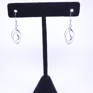 Pode incluir: Um par de brincos pendentes em tom prateado com um design de espiral de hélice dupla. Cada brinco consiste em duas fitas metálicas entrelaçadas que formam uma estrutura aberta e torcida, suspensa por um gancho de prata simples.