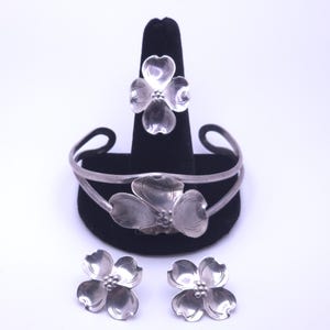 Può includere: Un set di gioielli in argento composto da un anello, un bracciale rigido e un paio di orecchini a lobo. Ogni pezzo presenta un design a fiore con quattro petali e un gruppo centrale di piccole sfere. Il metallo ha una finitura lucida.