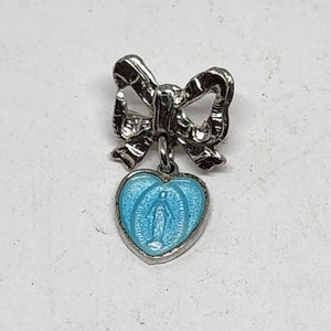 May include: A silver charm with a blue enamel heart featuring a religious figure. The charm is attached to a silver bow.