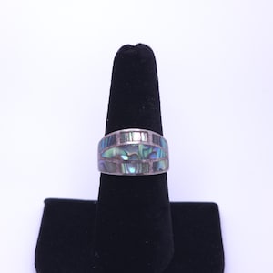 May include: A silver ring featuring a wide band with an inlay of iridescent abalone shell pieces. The shell segments are arranged in a geometric pattern with rectangular and curved shapes, displaying shades of green, blue, and purple throughout.