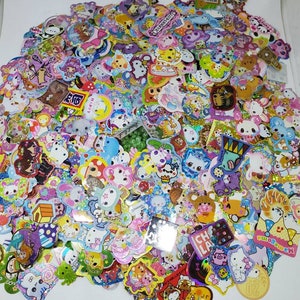May include: A large assortment of colorful stickers featuring various cartoon characters and designs. The stickers include images of animals, food, and other cute illustrations, with a variety of shapes and sizes. Many stickers have glitter accents.
