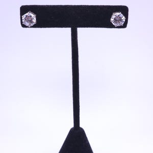 Puede incluir: Un par de pendientes de botón redondos, transparentes y facetados, engastados en garras de metal plateado. Los pendientes se muestran en un expositor de joyas en forma de T de terciopelo negro, resaltando el brillo de las piedras.