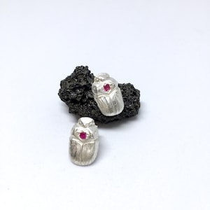 May include: A pair of silver earrings shaped like scarab beetles with a pink gemstone in the center of each beetle.
