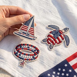 Op de afbeelding: Drie geborduurde patches met patriottische thema's: een zeilboot met een Amerikaanse vlag als zeil, een zeeschildpad met een schild in vlagpatroon en een kralenarmband-ontwerp met een sterbedel, uitgevoerd in de kleuren rood, wit en blauw.