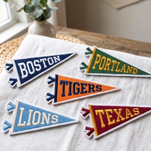 May include: Five triangular felt pennants featuring embroidered lettering. The pennants are navy blue with white text reading BOSTON, green with yellow text reading PORTLAND, orange with navy text reading TIGERS, light blue with cream text reading LIONS, and maroon with yellow text reading TEXAS.