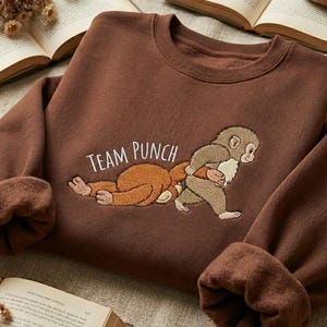 May include: A brown sweatshirt with the embroidered text "TEAM PUNCH" and a cartoon monkey design. The design features two monkeys, one lying down and the other standing. The sweatshirt has a crew neck and long sleeves with ribbed cuffs.