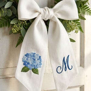 Op de afbeelding: Een witte linnen strik met een blauwe geborduurde hortensia en de letter "M". De strik maakt deel uit van een krans met groen blad. De strik hangt aan een witte deur.