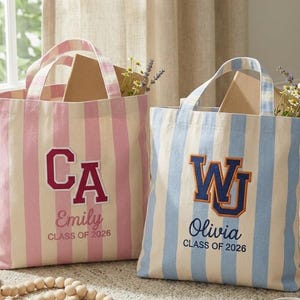 May include: Two striped tote bags. One is pink and cream with "CA" and "Emily, Class of 2026". The other is blue and cream with "WU" and "Olivia, Class of 2026". Both have handles and are filled with items.
