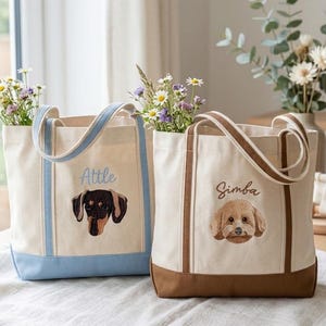 Pode incluir: Duas sacolas de lona com retratos de cães bordados e arranjos florais. Uma sacola é azul claro com o nome "Attle" e um dachshund. A outra é marrom com o nome "Simba" e um poodle.