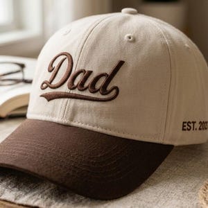 Może przedstawiać: Beżowa czapka bejsbolowa z brązowym daszkiem i napisem "Dad" wyhaftowanym brązową czcionką. Tekst "EST. 2013" jest wyhaftowany z boku. Czapka leży na powierzchni w neutralnym kolorze.