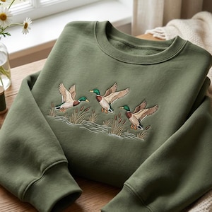 Puede incluir: Una sudadera verde oliva con un dise&ntilde;o bordado de tres patos en vuelo sobre juncos y agua. La sudadera tiene cuello redondo y pu&ntilde;os acanalados. El dise&ntilde;o est&aacute; detallado con colores marr&oacute;n, verde y beige.