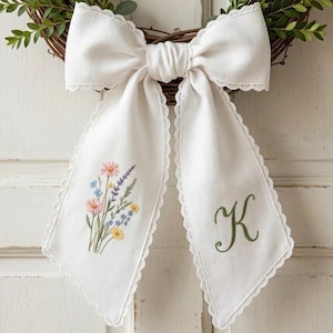May include: A white fabric bow with floral embroidery and a green initial "K" on the tails, attached to a grapevine wreath with green foliage. The bow has a delicate lace trim.