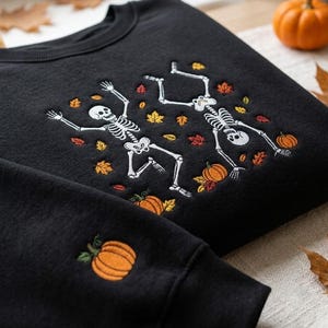 Op de afbeelding: Een zwarte sweater met ronde hals voorzien van een geborduurd ontwerp van twee witte skeletten die dansen tussen verspreide oranje, gele en rode herfstbladeren en kleine oranje pompoenen. Een extra kleine oranje pompoen is op de mouw geborduurd.
