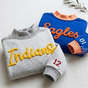 May include: A gray sweatshirt with a yellow chenille 'Indians' script and the number '12' on the sleeve, paired with a blue sweatshirt featuring an orange 'Eagles' script and the number '01' on the sleeve, both with gingham collars and cuffs.