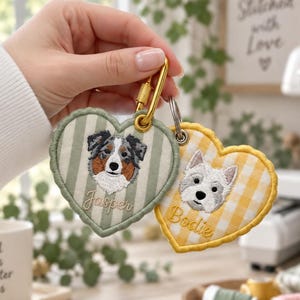 Puede incluir: Dos llaveros bordados en forma de corazón con retratos de perros. Uno tiene rayas verdes con un pastor australiano y el nombre Jasper. El otro tiene un patrón de cuadros amarillos con un terrier blanco y el nombre Bodie.