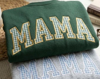 Op de afbeelding: Twee sweatshirts, één bosgroen en één grijs, zijn op elkaar gestapeld. Het groene sweatshirt heeft het woord "MAMA" geborduurd in een ruitpatroon met gouden rand. Het grijze sweatshirt heeft hetzelfde woord geborduurd in een blauw ruitpatroon.