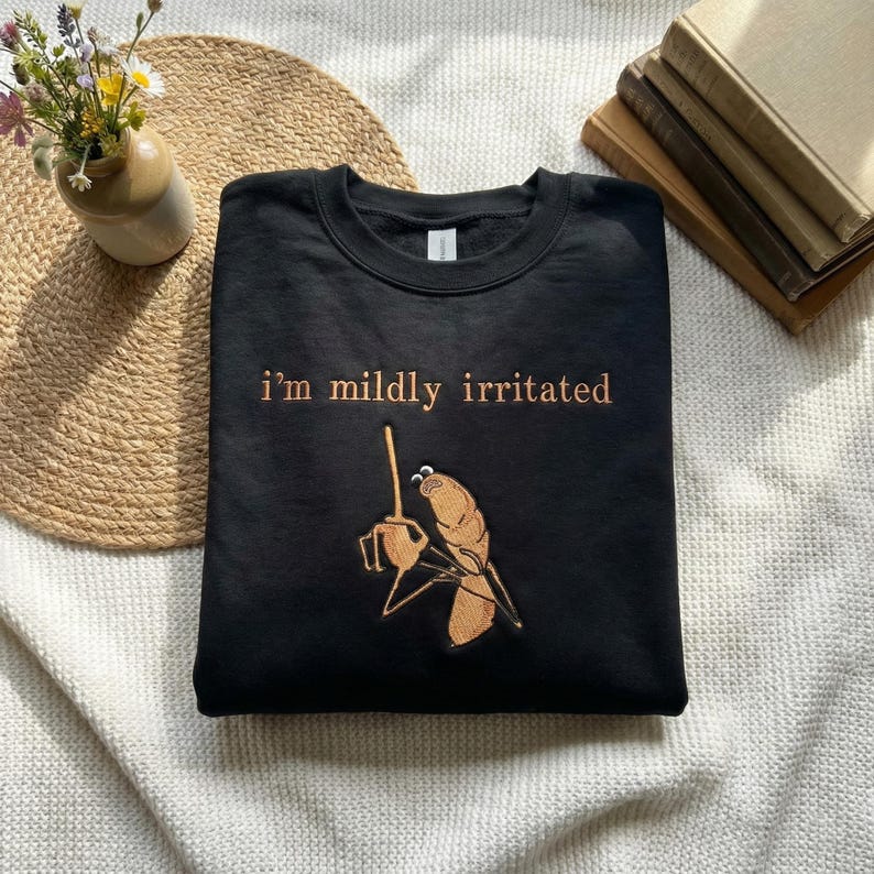 Może przedstawiać: Czarna bluza z haftowanym napisem "I'm mildly irritated" w kolorze miedzianym. Poniżej tekstu znajduje się haftowany wz&oacute;r postaci z kresk&oacute;wki trzymającej filiżankę kawy. Bluza jest złożona na białej powierzchni.