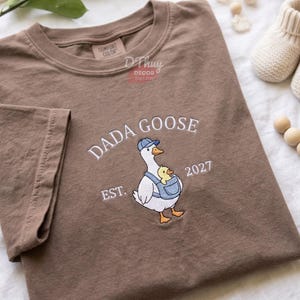 May include: A brown short-sleeve t-shirt featuring an embroidered graphic of a white goose wearing a blue cap and carrying a yellow chick in a blue baby carrier. The text reads DADA GOOSE EST. 2027 in white thread. The shirt has a crew neckline.