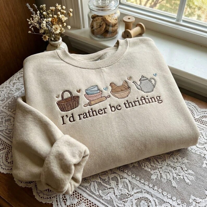 May include: Beige sweatshirt with embroidered kitchen-themed graphics, including a basket, bowls, rolling pin, chicken, and teapot. The text "I'd rather be thrifting" is embroidered below the graphics. The sweatshirt is folded on a lace tablecloth.
