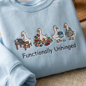 Könnte beinhalten: Hellblaues Sweatshirt mit einer Stickerei, die vier Cartoon-Gänse in verschiedenen Szenarien zeigt. Eine Gans arbeitet an einem Laptop, eine andere spielt mit Spielzeug, eine dritte hält einen Knetbehälter. Der Text "Functionally Unhinged" ist darunter gestickt.