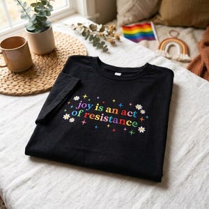 Pu&ograve; includere: T-shirt nero con la scritta ricamata "joy is an act of resistance" in lettere colorate e dettagli floreali. La maglietta &egrave; piegata su una superficie bianca, con una tazza e una pianta sullo sfondo.