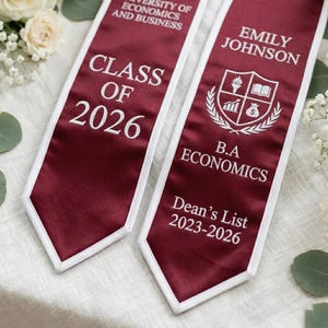 May include: Maroon graduation sash with white trim. Text includes "CLASS OF 2026", "EMILY JOHNSON", "B.A ECONOMICS", and "Dean's List 2023-2026". The sash features a university crest with a book, flame, and money bag.