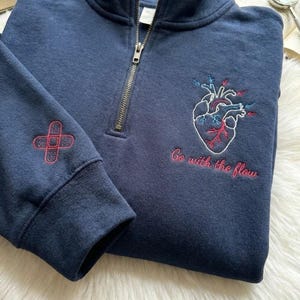 Könnte beinhalten: Ein marineblaues Sweatshirt mit einem Viertel-Reißverschluss-Ausschnitt. Das Sweatshirt hat ein gesticktes anatomisches Herzdesign mit roten und blauen Akzenten und den Schriftzug "Go with the flow". Auf dem Ärmel ist ein rotes Kreuzdesign gestickt.