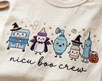 Może przedstawiać: Kremowy t-shirt z wyhaftowanymi postaciami sprzętu medycznego w stylu halloweenowym, w tym butelką dla niemowląt wampirem i strzykawką z wiaderkiem w kształcie dyni. Napis "nicu boo crew" jest wyhaftowany czarną kursywą poniżej.