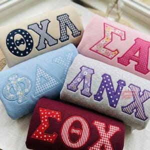 Peut inclure: Cinq sweat-shirts pliés avec de grandes lettres grecques en appliqué. Le sweat-shirt crème présente des lettres bleu marine à étoiles et vichy. Le rose a des lettres à fleurs et à pois roses. Le bleu montre des lettres bleu clair à fleurs et vichy. Le gris présente des lettres violettes à fleurs, à pois et rayées. Le sweat-shirt bordeaux a des lettres rouges à étoiles, rayées et vichy.