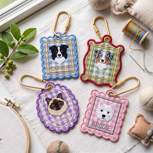 May include: Four embroidered pet portrait keychains with gold-tone carabiners. Each features a scalloped frame on a gingham background: a blue frame with a black and white dog named CHARLIE, a red frame with a tri-color dog named LUNA, a purple oval frame with a Siamese cat named WILLOW, and a pink frame with a white dog named BRODIE.
