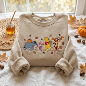 May include: A beige crewneck sweatshirt featuring an embroidered design of Eeyore carrying a pumpkin, Piglet, Winnie the Pooh holding a honey pot, and Tigger walking together. The scene is decorated with scattered autumn leaves and acorns.