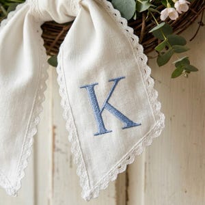 May include: Cream-coloured linen scarf featuring a light blue embroidered letter "K" and a scalloped lace trim. The scarf is tied in a bow, with a wreath of foliage and flowers in the background.