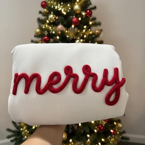 Może przedstawiać: Biała prostokątna poduszka z napisem "merry" w czerwonych, kursywnych literach. Poduszka jest trzymana przed choinką ze złotymi i czerwonymi ozdobami.
