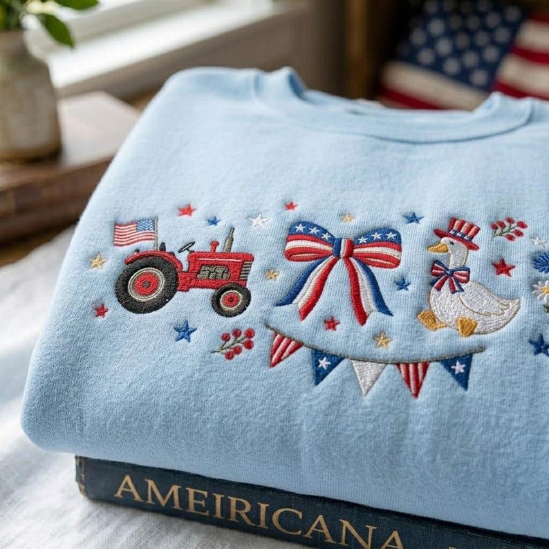 K&ouml;nnte beinhalten: Hellblaues Sweatshirt mit gestickten patriotischen Designs. Es zeigt einen roten Traktor mit amerikanischer Flagge, eine Schleife, eine Ente mit Zylinder und Stern- und Bannerakzente. Das Sweatshirt liegt gefaltet auf einem Buch.