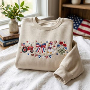 K&ouml;nnte beinhalten: Beiges Sweatshirt mit gesticktem patriotischem Design. Das Design umfasst einen roten Traktor, eine Schleife mit rot-wei&szlig;-blauen Streifen, eine Gans mit Zylinder und verschiedene Blumen und Sterne. Das Sweatshirt hat einen Rundhalsausschnitt und lange &Auml;rmel.
